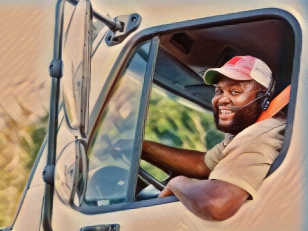 A truck driver looking at the camera as he sits in the driver's seat, arm hanging out the open window. He is an African-American man in his 30s with a large build and a smile on his face.